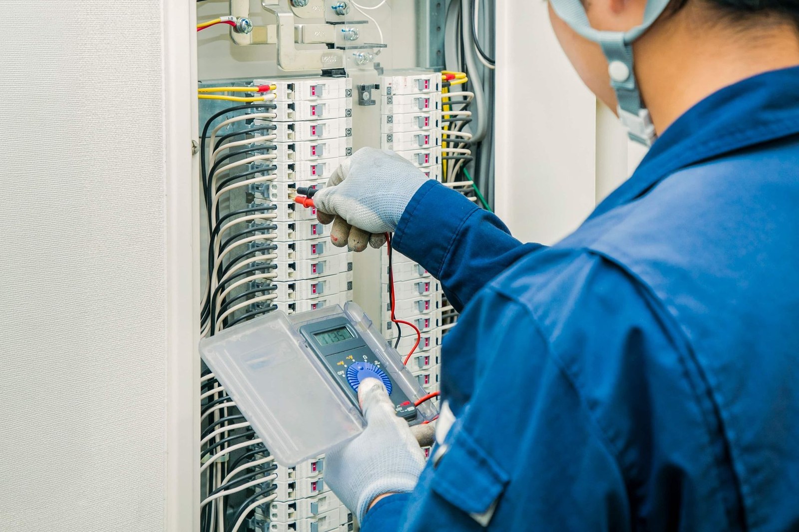 Technician in a blue uniform inspecting wiring in an electrical control panel with a tablet